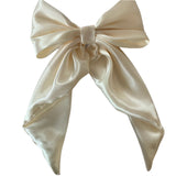 A cream satin hair bow clip with long tails, attached to a silver-colored barrette clip, displayed on a white background.
