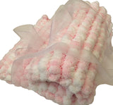 A hand knitted pink and white baby blanket with pom poms, tied with a ribbon.