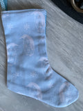 A light blue fabric Christmas stocking with a pale gray penguin and snowflake pattern, laid flat on a gray textured surface.