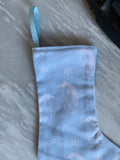 A light blue fabric Christmas stocking with a penguin and snowflake pattern, featuring a matching blue patterned hanging loop, shown on a gray marble surface.