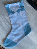A light blue Christmas stocking with white snowflake and penguin patterns, featuring a large white heart with the number 6 on the cuff, laid flat on a gray textured surface.