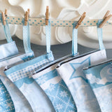 A close-up of several light blue and white fabric Christmas stockings hanging from a  ribbon with small wooden pegs. The stockings feature various holiday patterns including snowflakes, stars, gingham, and moons, set against a decorative white background.