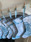 A group of light blue Christmas stockings with various festive patterns, hanging from a wooden dowel with small wooden pegs..