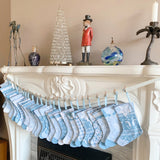 A set of light blue and white patterned Christmas stockings hanging from a white fireplace mantel with wooden clips. The mantel is decorated with a crystal tree, a nutcracker, a blue vase, and a palm candle holder against a beige wall.