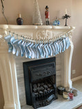 A string of light blue and white cotton fabric Christmas stockings hanging across an ornate white fireplace mantel, with candles, decorative bottles, and a nutcracker displayed above.