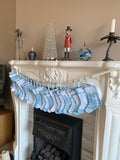 A string of light blue and white patterned Christmas stockings hanging across a white fireplace mantel, with holiday decor including a crystal tree, figurines, and ornaments in the background.