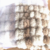 A close-up of a textured, chunky knit blanket pram cover in cream, beige, and light brown colours, with a sheer white mesh ribbon on one side.