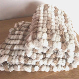 A chunky knit blanket with alternating thick beige, brown and off-white pom poms, folded on a wooden tabletop against a plain light background.