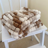 Textured blanket with pom-pom pattern on a white stool against a neutral wall.