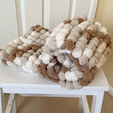 Knitted blanket with brown, beige, and white pom-poms on a white chair.
