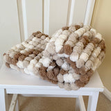 Knitted blanket with brown, beige, and white pom-poms on a white chair.