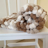 Knitted blanket with brown, beige, and white pom-poms on a white chair.