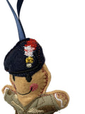 A close-up of a handmade embroidered felt ornament shaped like a soldier, wearing a dark navy blue beret with red, white, and gold embroidery, a beige face, and a green camouflage jacket, hanging from a blue ribbon against a plain light background.