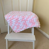A chunky, textured blanket pram cover in pink and white yarn is draped over a small white wooden stool in a home setting with beige carpet and a cream paneled door.