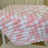 A chunky knit blanket with alternating fluffy pink and white loops, draped over a white chair in front of a beige paneled wall.