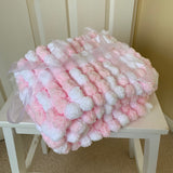 A fluffy pink and white textured blanket with pom-pom details, tied with a sheer white ribbon, resting on a small white stool in front of a cream paneled door.