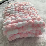 A folded plush blanket with a textured bumpy pattern in alternating pink and white loops, resting on a soft gray blanket.