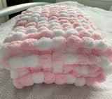 A folded plush chenille blanket with a pink and white pom-pom striped pattern, resting on a textured light gray blanket.