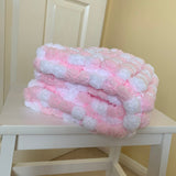 A folded plush blanket cover with alternating pink and white textured squares sits on a white wooden chair in a home setting.