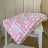 A chunky, textured knit blanket in pink and white is draped over a white wooden chair in front of a cream-colored paneled door.