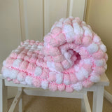 A pink and white chunky pom-pom blanket cover rolled on a white wooden stool in front of a beige paneled door.
