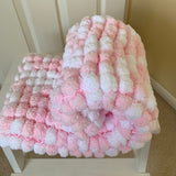 A chunky, blanket cover in pink and white, draped over a white wooden chair in an indoor setting.