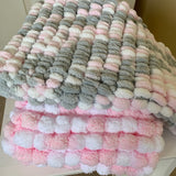 Two plush textured throw blankets stacked on a white table. The top blanket features a striped pattern of gray, white, and light pink loops.