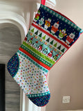 A festive Christmas stocking with a colorful pattern of cartoon animals, snowmen, penguins, and Christmas trees, hanging from a red fabric loop in front of a white fireplace mantel.