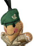 A close-up of a handmade felt ornament shaped like a smiling soldier with a green beret, embroidered facial features, and a camouflage-patterned body, hanging from a black ribbon against a plain light background.