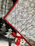 A close-up of the back of a quilted fabric advent calendar showing pockets for dowel, featuring a gray and white snowflake pattern, bordered by red and white polka dot trim, layered over other holiday-themed quilts with snowmen, trees, and plaid designs.