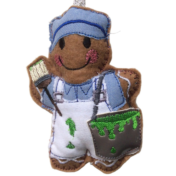 Gingerbread man decoration wearing a blue painter's outfit with green paint on his overalls, holding a paintbrush. On white background.