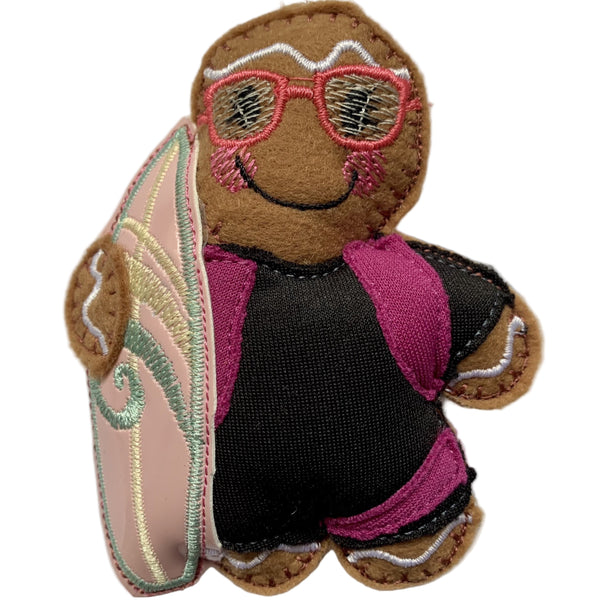 Surfer Girl Gingerbread Christmas Decoration. Dressed in a black wetsuit with pink detail,  holding a pink surfboard. On white background.